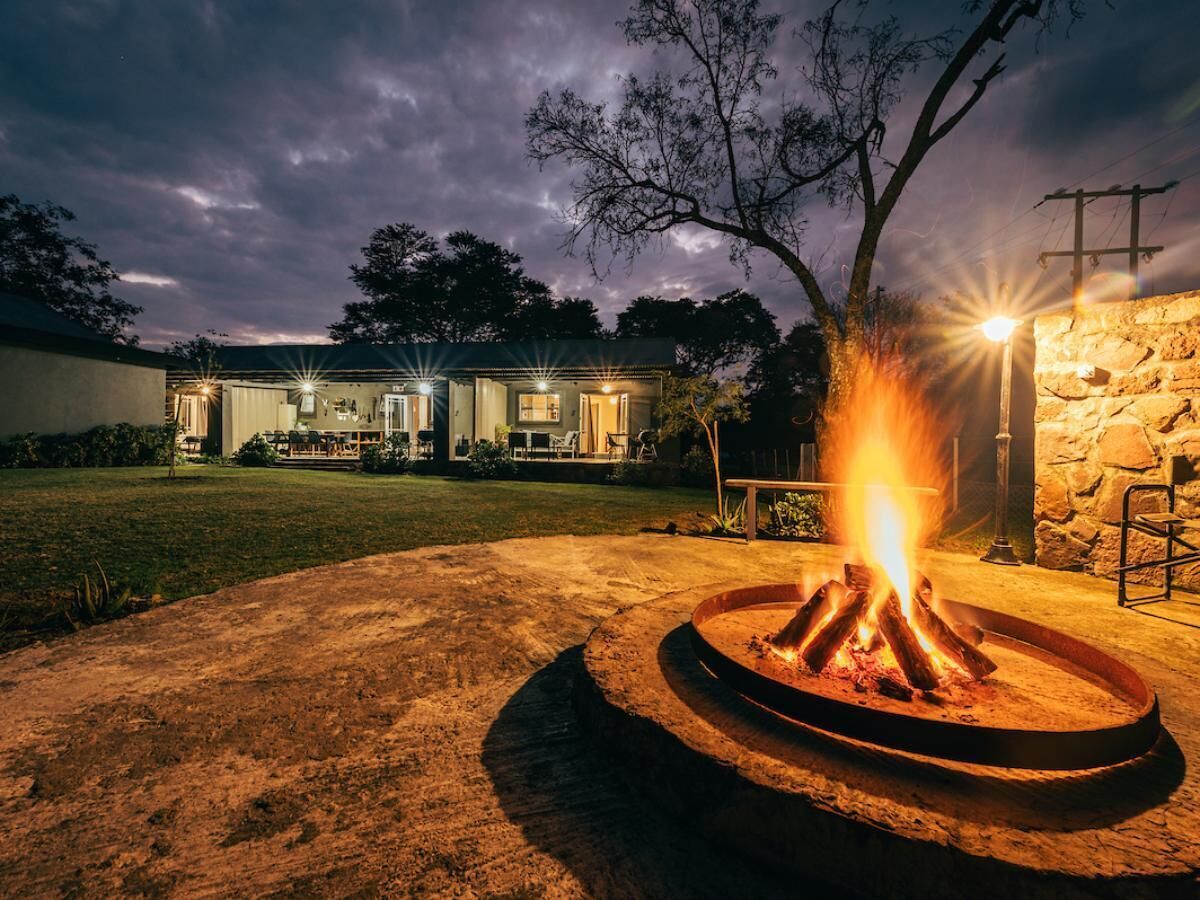 A fire pit in a circular metal frame with logs, surrounded by a dirt area, with a house and stone wall in the background.