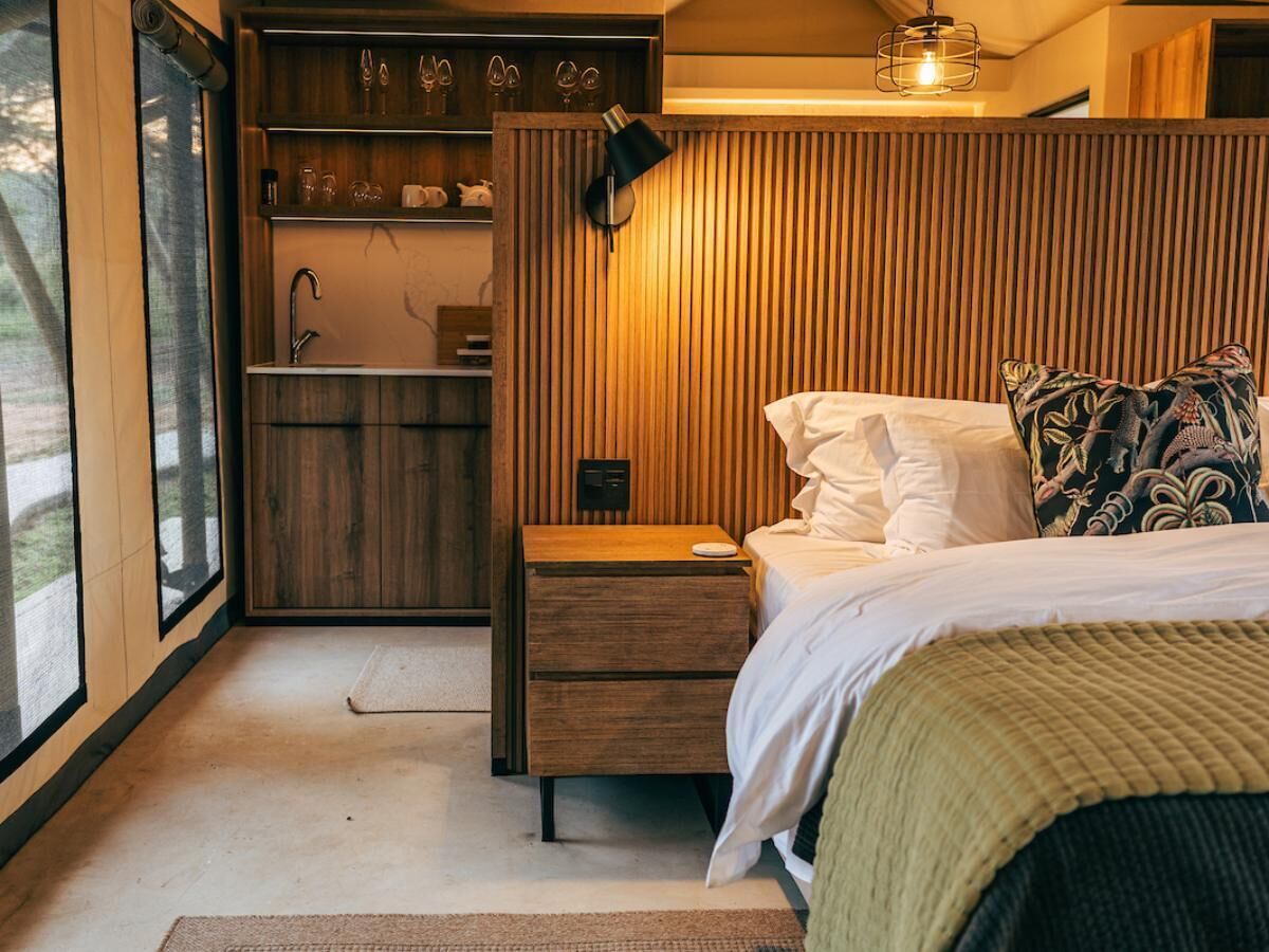 A bedroom with a wooden headboard, a bedside table, a bed with white bedding, and a window with a view of the outdoors.