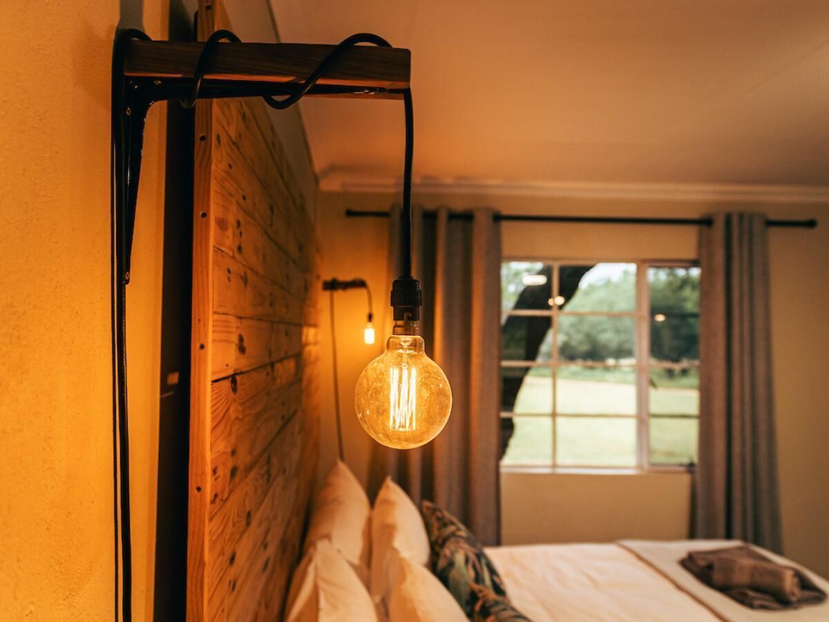 A bedroom with a wooden headboard, a hanging light bulb, a window with curtains, and a view of greenery outside.