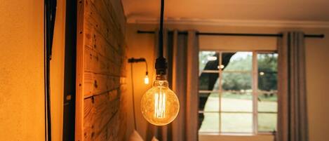 A bedroom with a wooden headboard, a hanging light bulb, a window with curtains, and a view of greenery outside.