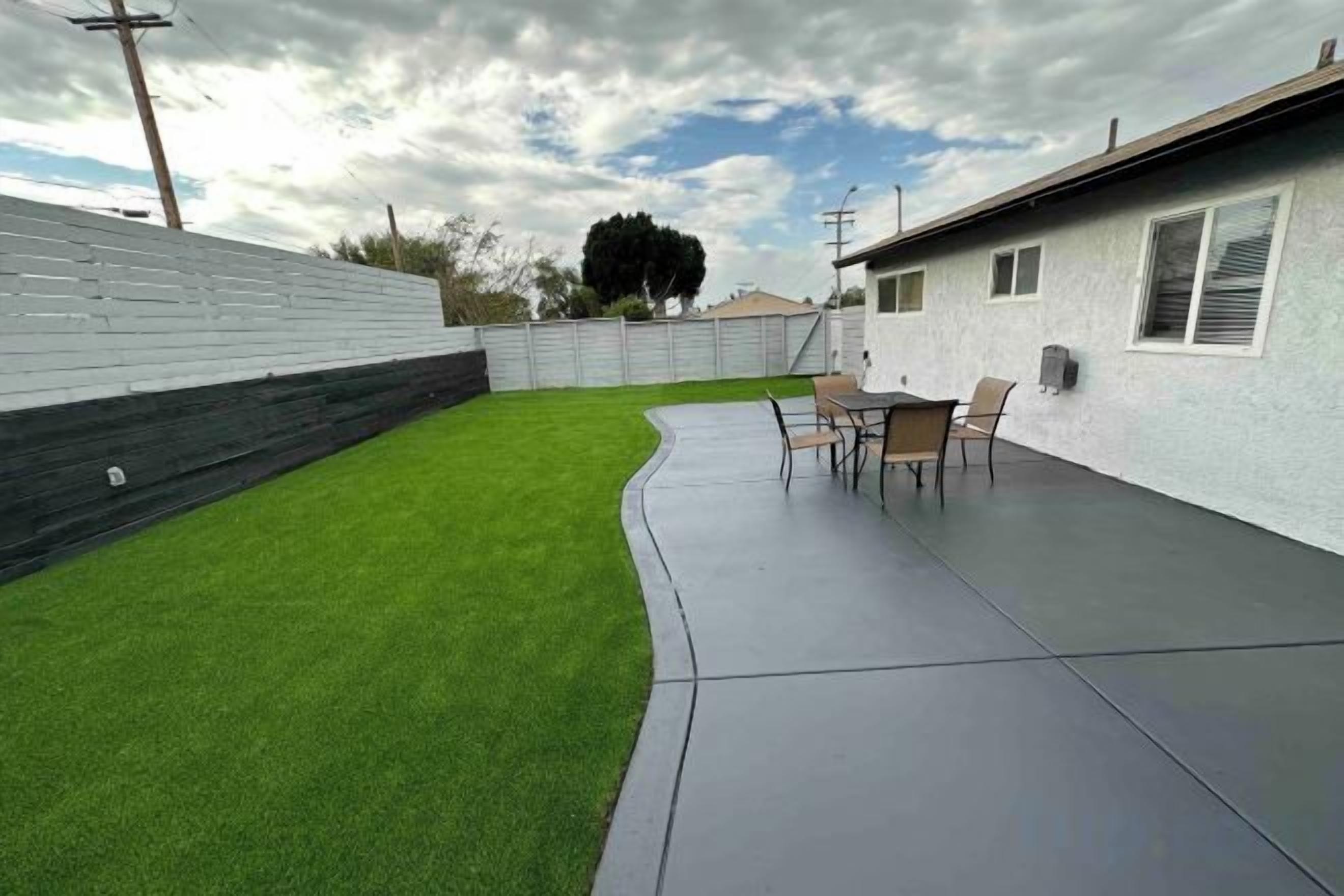 A backyard with artificial grass, a patio with a table and chairs, and a white building with windows.