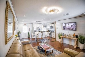 A spacious living area with a sofa, coffee table, and a dining area with chairs. There is a kitchen in the background with modern appliances and a large TV mounted on the wall. - Xlnc Exotic Home Sunnyside by sdstay (San Diego)