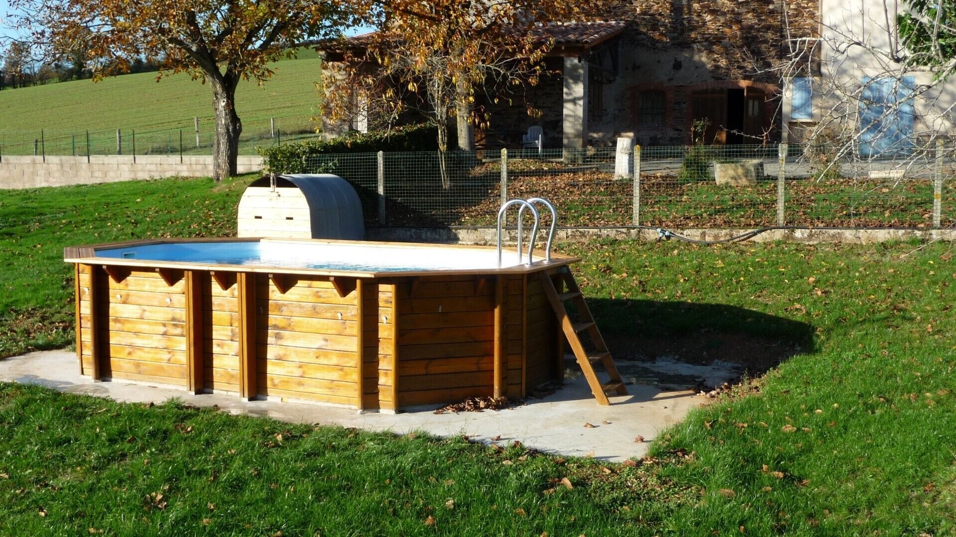A wooden hot tub with a ladder, surrounded by a grassy area and a stone building in the background.