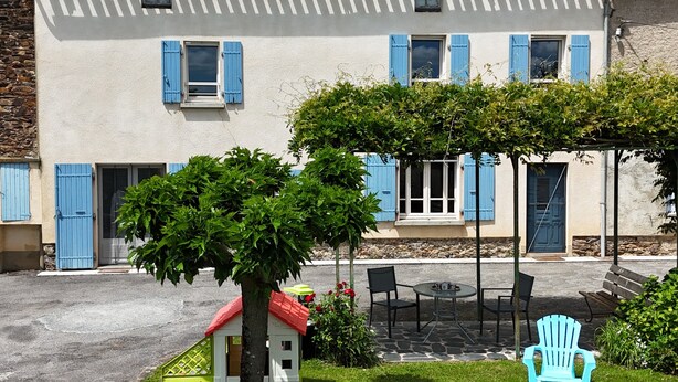 Un bâtiment avec des volets bleus, un petit arbre, des sièges à l’extérieur et une structure blanche qui ressemble à une maison.