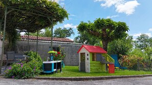 Ein kleines Spielhaus mit rotem Dach, einer grünen Spielkonstruktion und einem überdachten Sitzbereich mit Tisch und Stühlen.