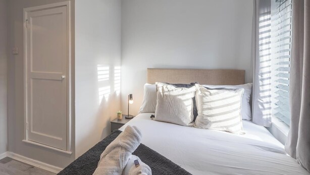Ein Schlafzimmer mit Bett, Kissen, einem Nachttisch, einer Lampe, einem Fenster mit Jalousien und einer Tür.