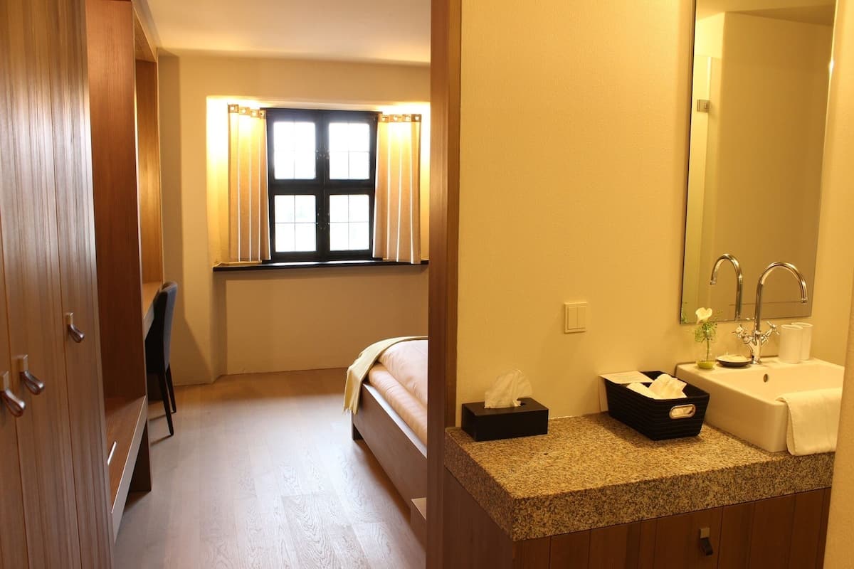 A hotel room with a bed, a small desk, a window with curtains, a sink with a mirror, and a trash bin.