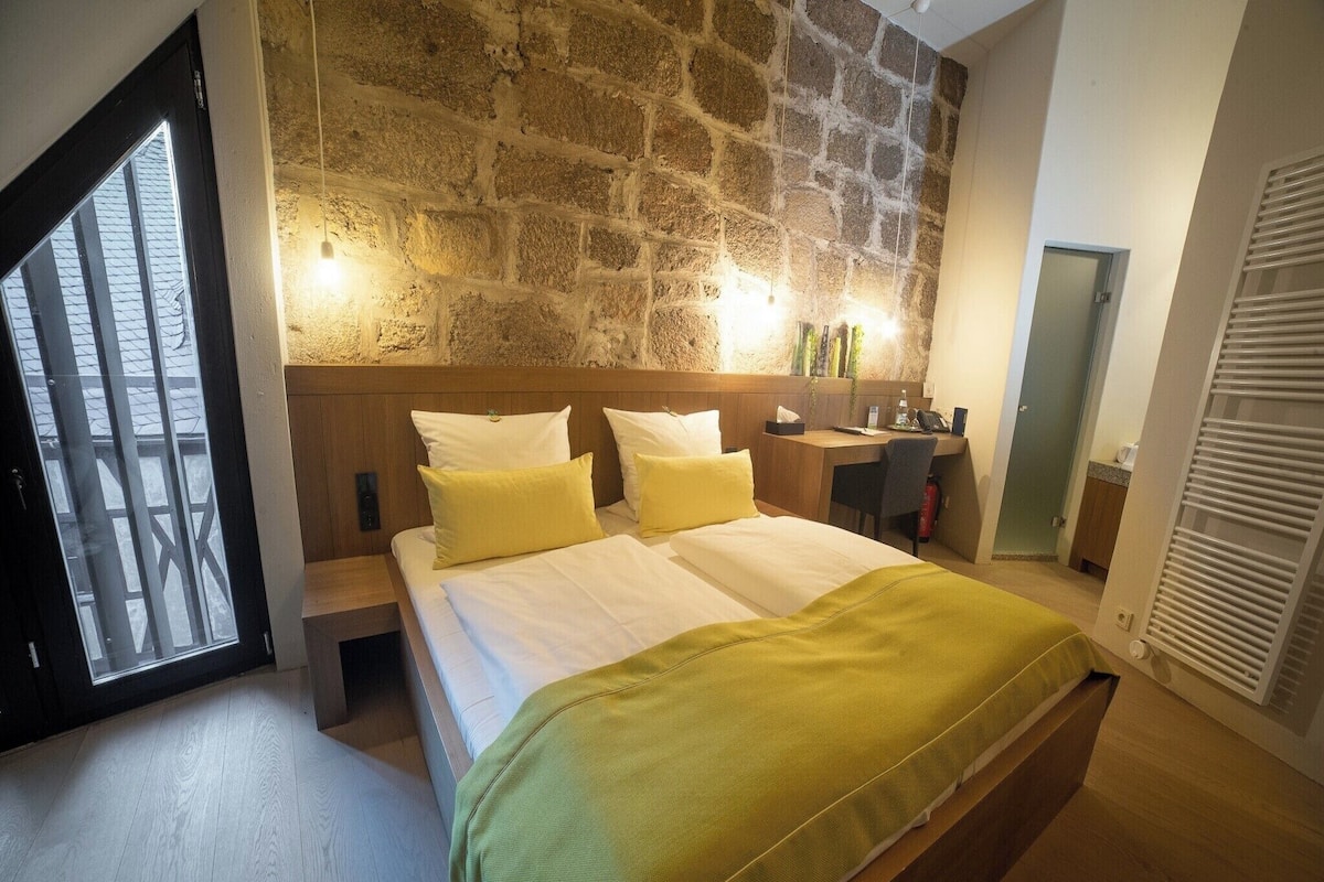 A bedroom with a stone wall, a bed with white and yellow bedding, a desk, and a mirror.