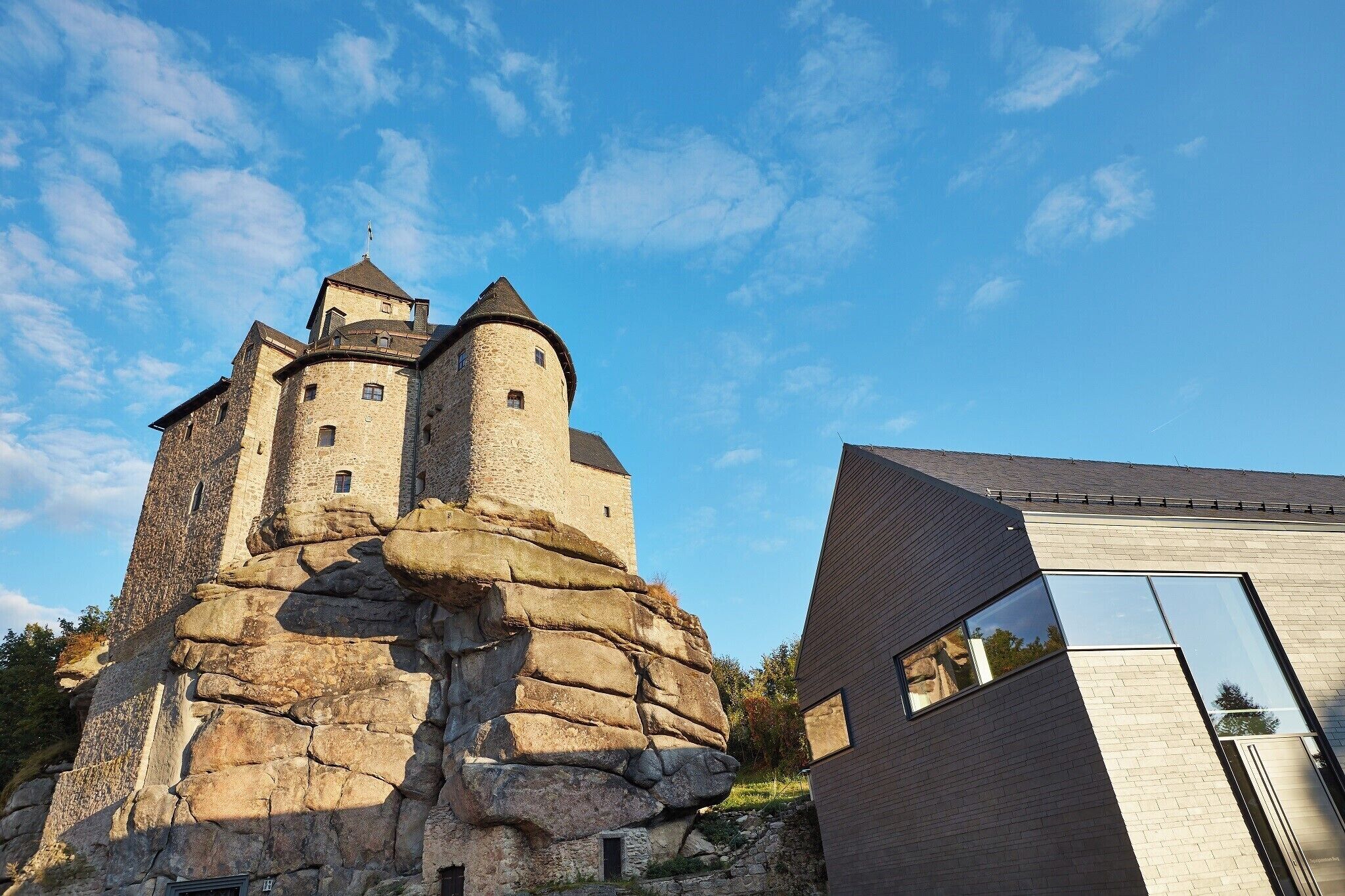 Un castillo de piedra histórico con una torre redonda, junto a un edificio moderno con grandes ventanales.
