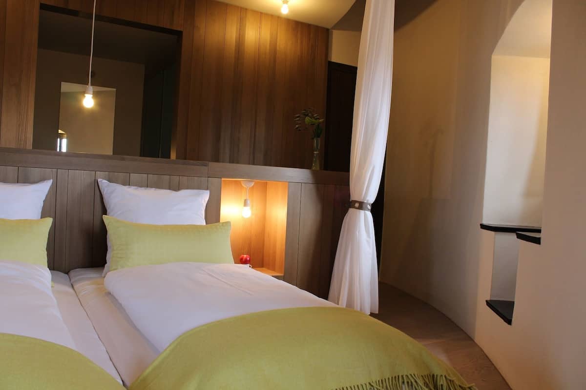 A hotel room with a wooden headboard, two beds with green bedding, a bedside table with a lamp, and a small plant.