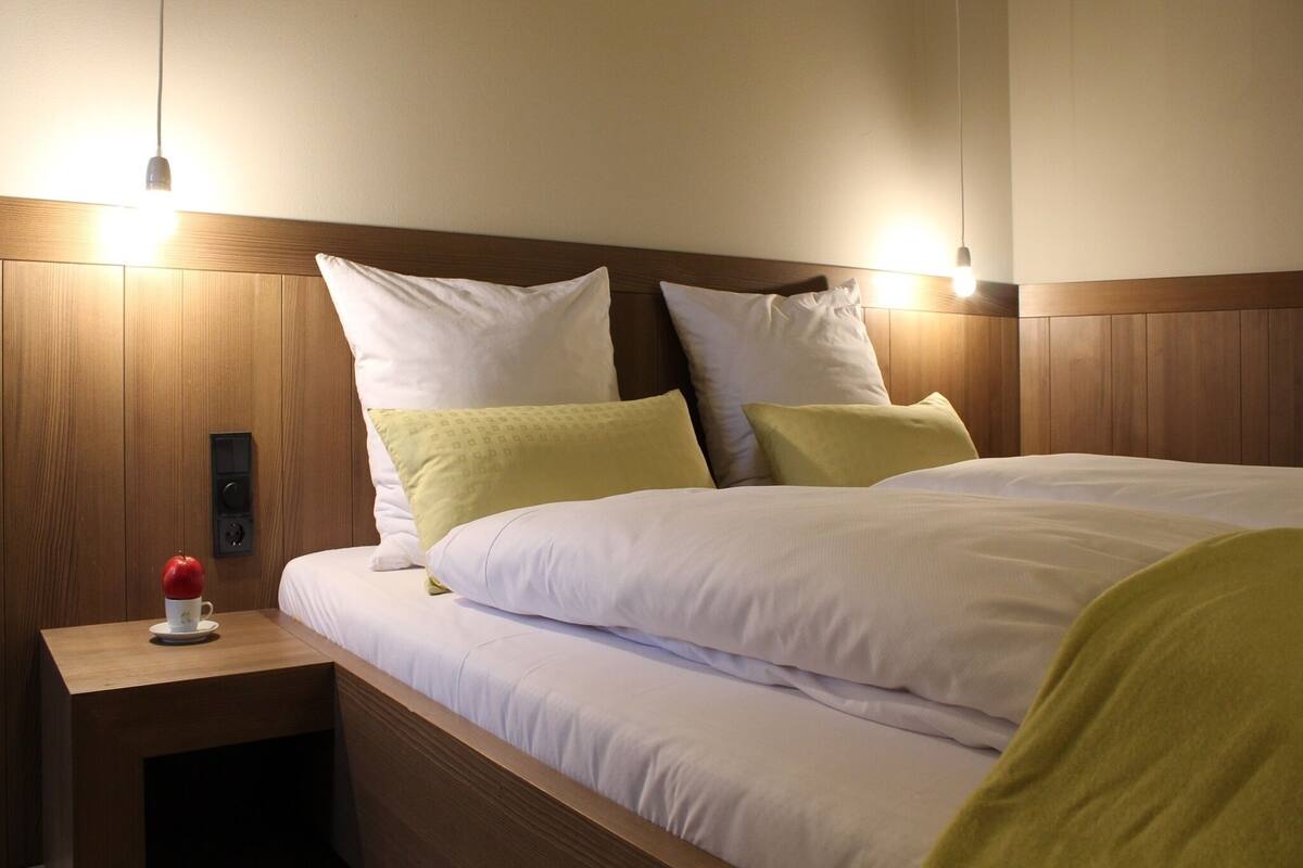 A neatly made bed with white linens and multiple pillows, a wooden nightstand with a red apple and a cup, and two wall-mounted lights.