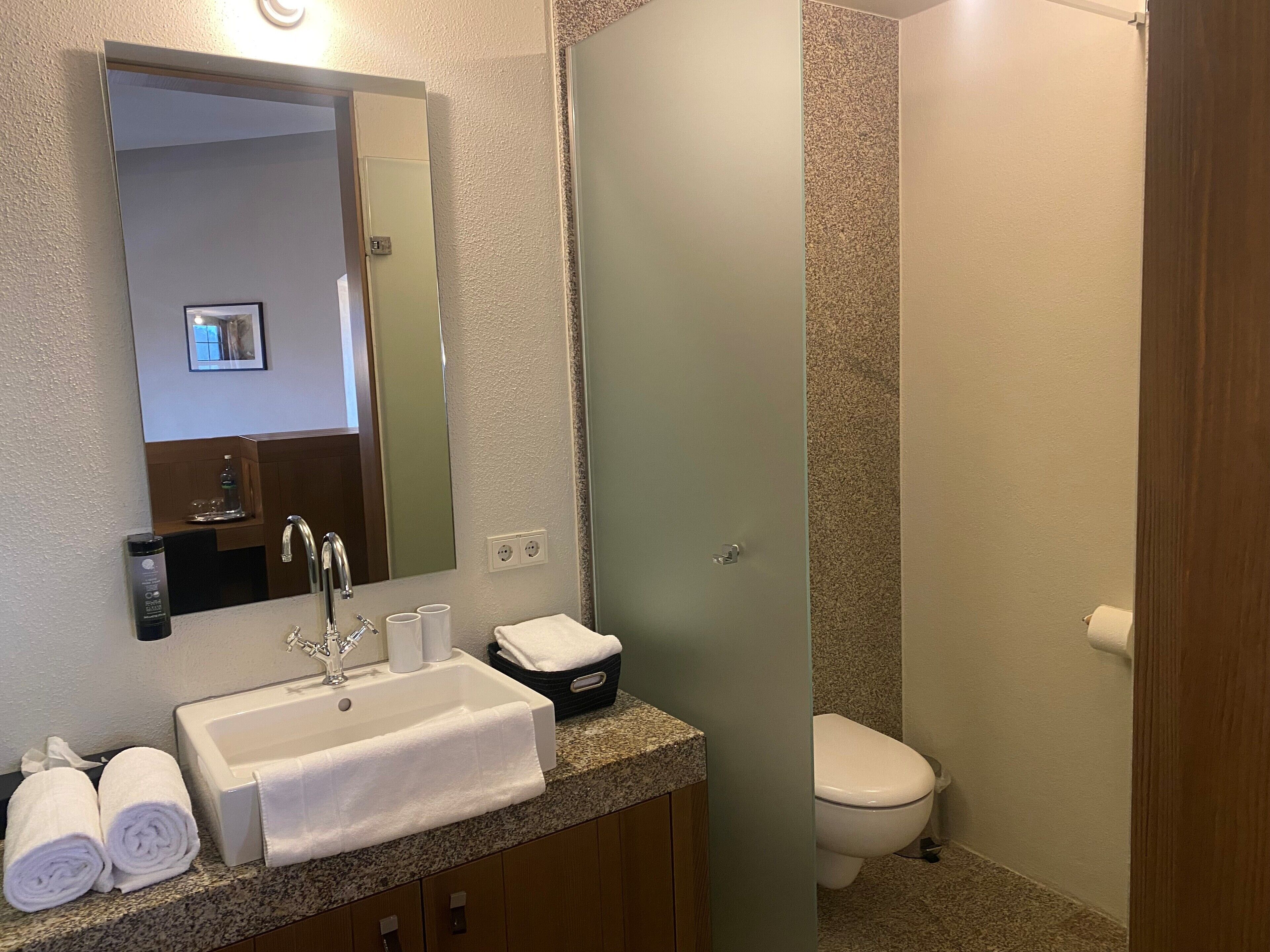 A bathroom with a white sink, mirror, and a toilet. There are towels and a bottle on the sink counter.