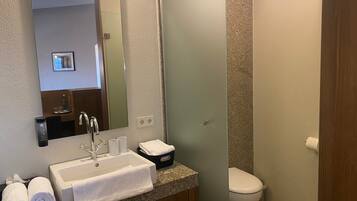 A bathroom with a white sink, mirror, and a toilet. There are towels and a bottle on the sink counter.