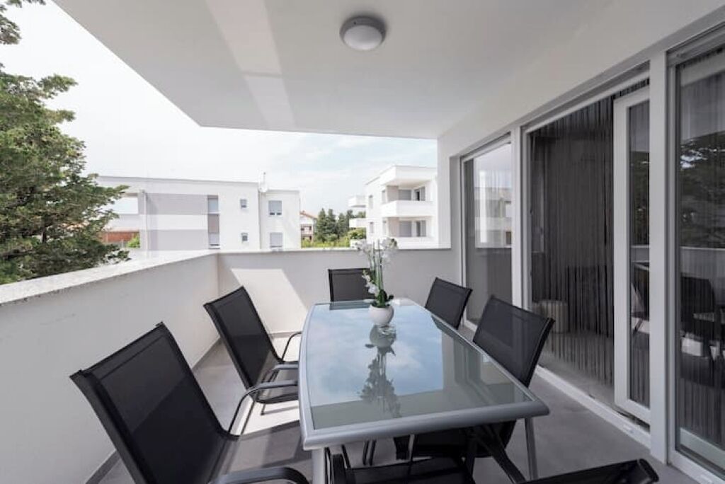 A modern balcony with a glass table and chairs, a laptop, and a vase with flowers.