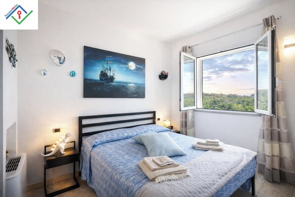 Un dormitorio con cama, mesita de noche, lámpara, ventana con vistas y un cuadro en la pared.