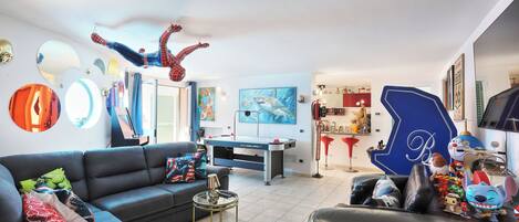 Una sala de estar con un sofá, una mesa de centro, una mesa de billar y varias decoraciones, incluyendo una figura de Spiderman, una silla azul y un cuadro en la pared.