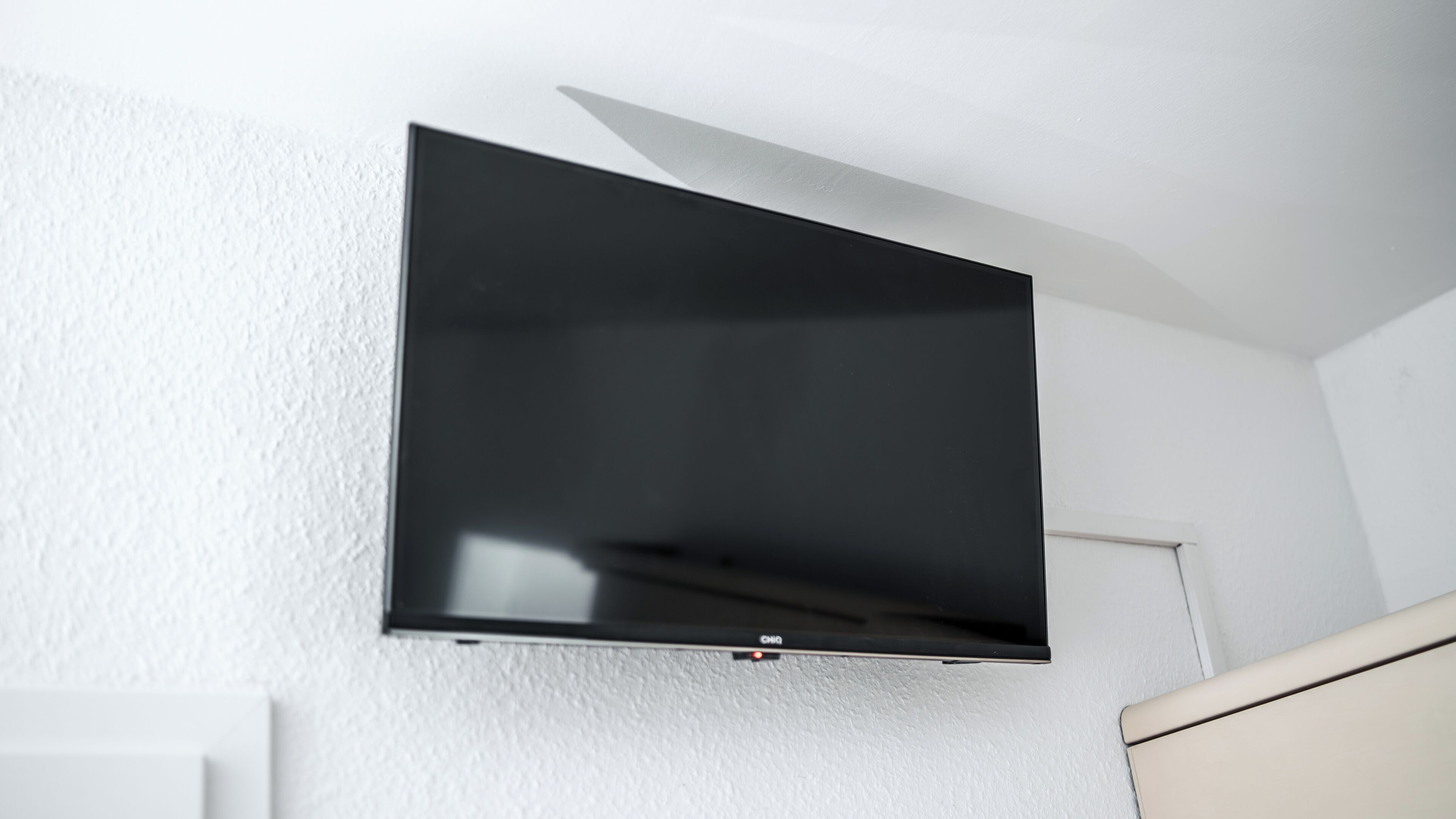 Un téléviseur à écran plat fixé au mur blanc d’une pièce où l’on trouve également une porte blanche et une commode blanche.