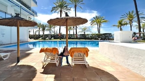 Ein Poolbereich mit Liegestühlen und Sonnenschirmen, ein Schwimmbecken, Palmen und ein Gebäude im Hintergrund.