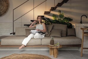 Una persona sentada en un sofá leyendo un libro, con una piña y bebidas sobre una mesita auxiliar, y una escalera al fondo.