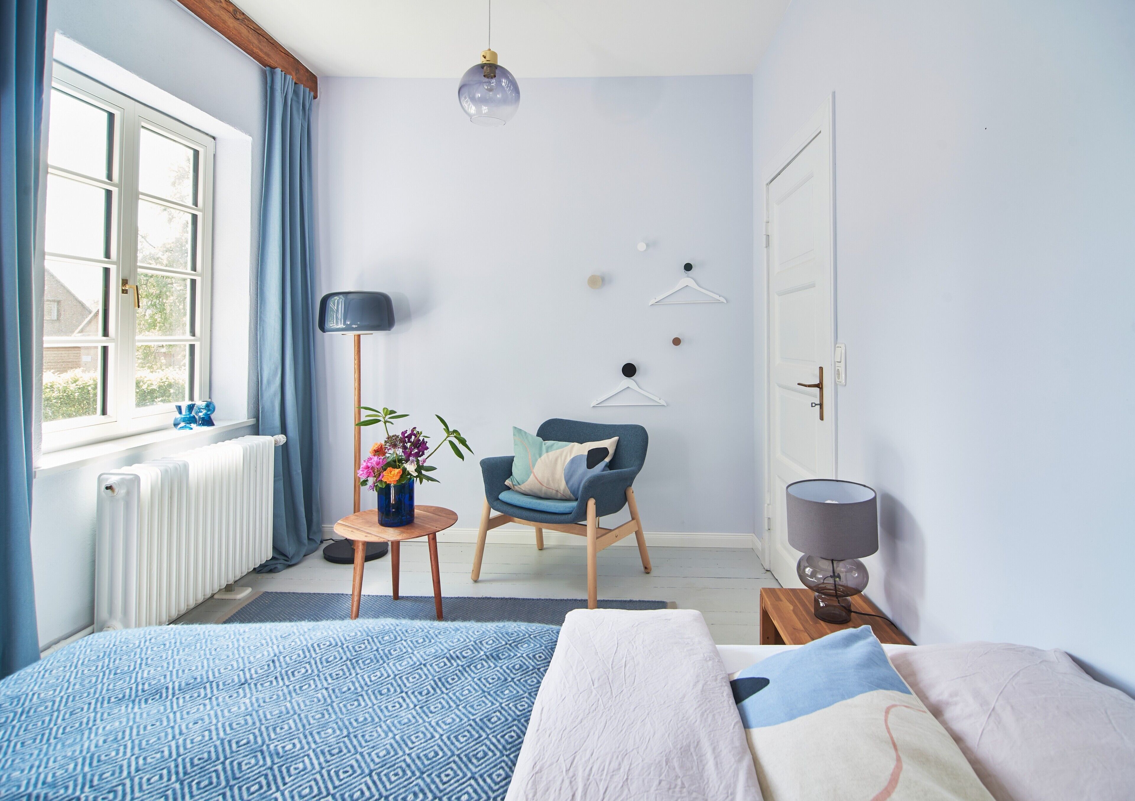 A bedroom with a bed, a chair, a small table with a vase of flowers, a floor lamp, and a radiator.