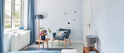 A bedroom with a bed, a chair, a small table with a vase of flowers, a floor lamp, and a radiator.