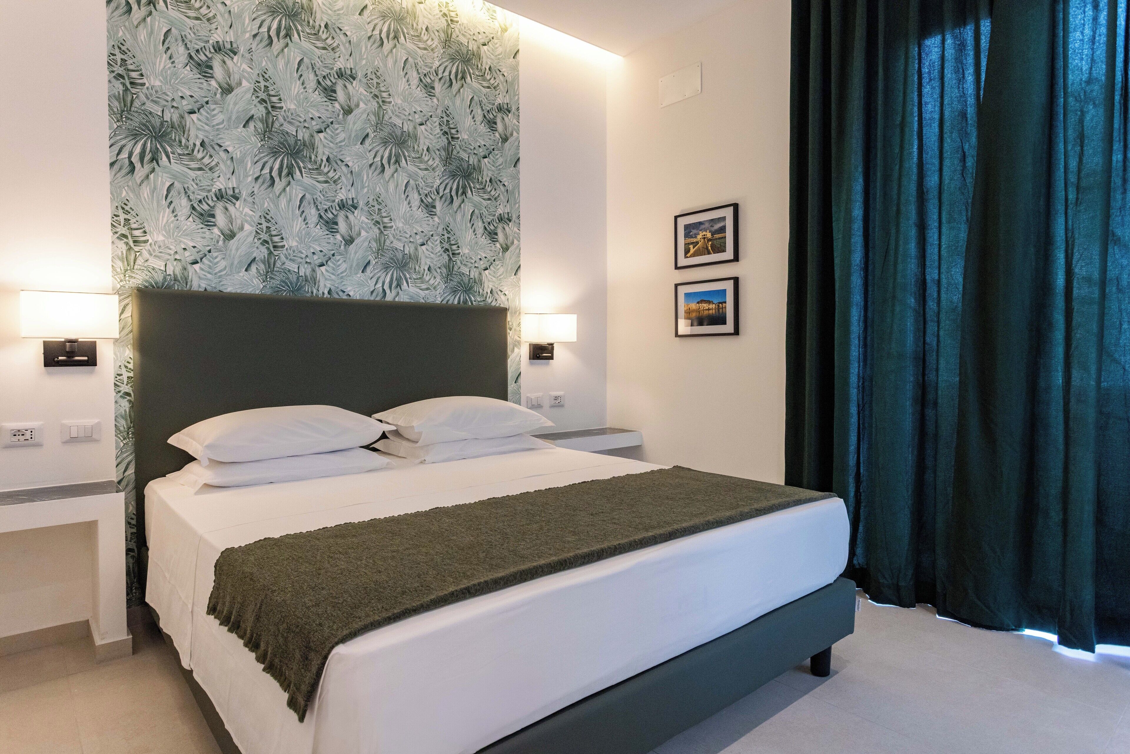 A neatly made bed with white linens and a dark green throw blanket, a headboard with a leaf-patterned wallpaper, two bedside lamps, and a dark curtain on the window.
