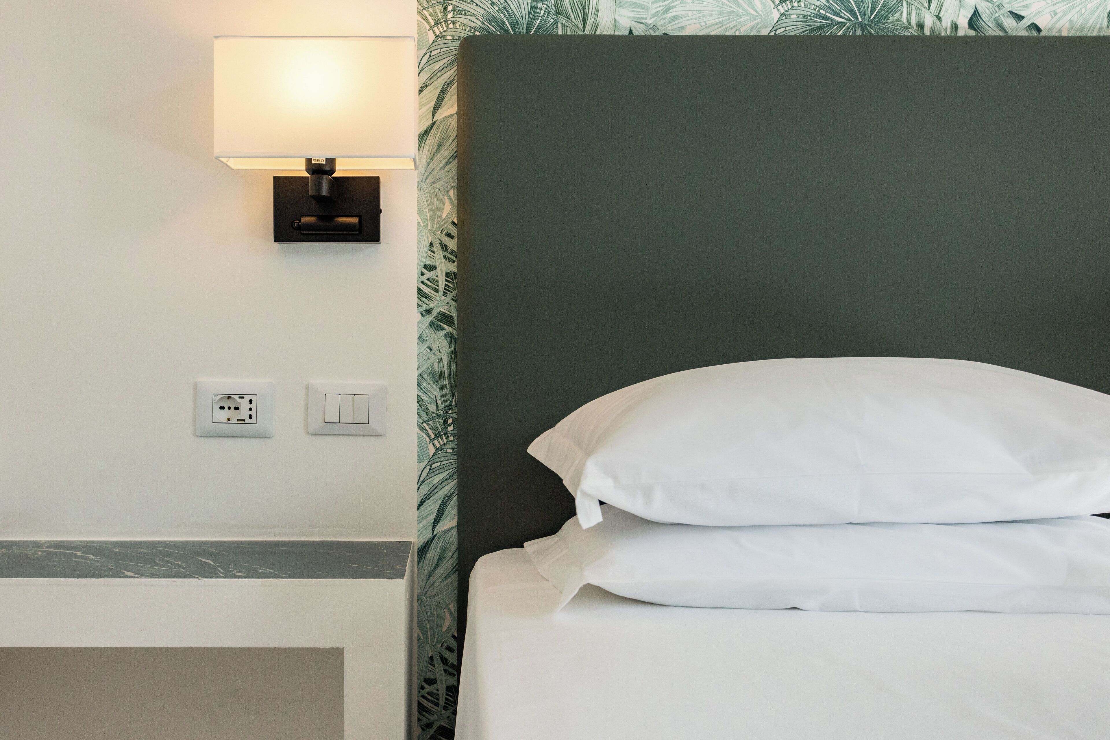 Habitació d'hotel amb un llit, una tauleta de nit, una làmpada penjada a la paret i una presa elèctrica.