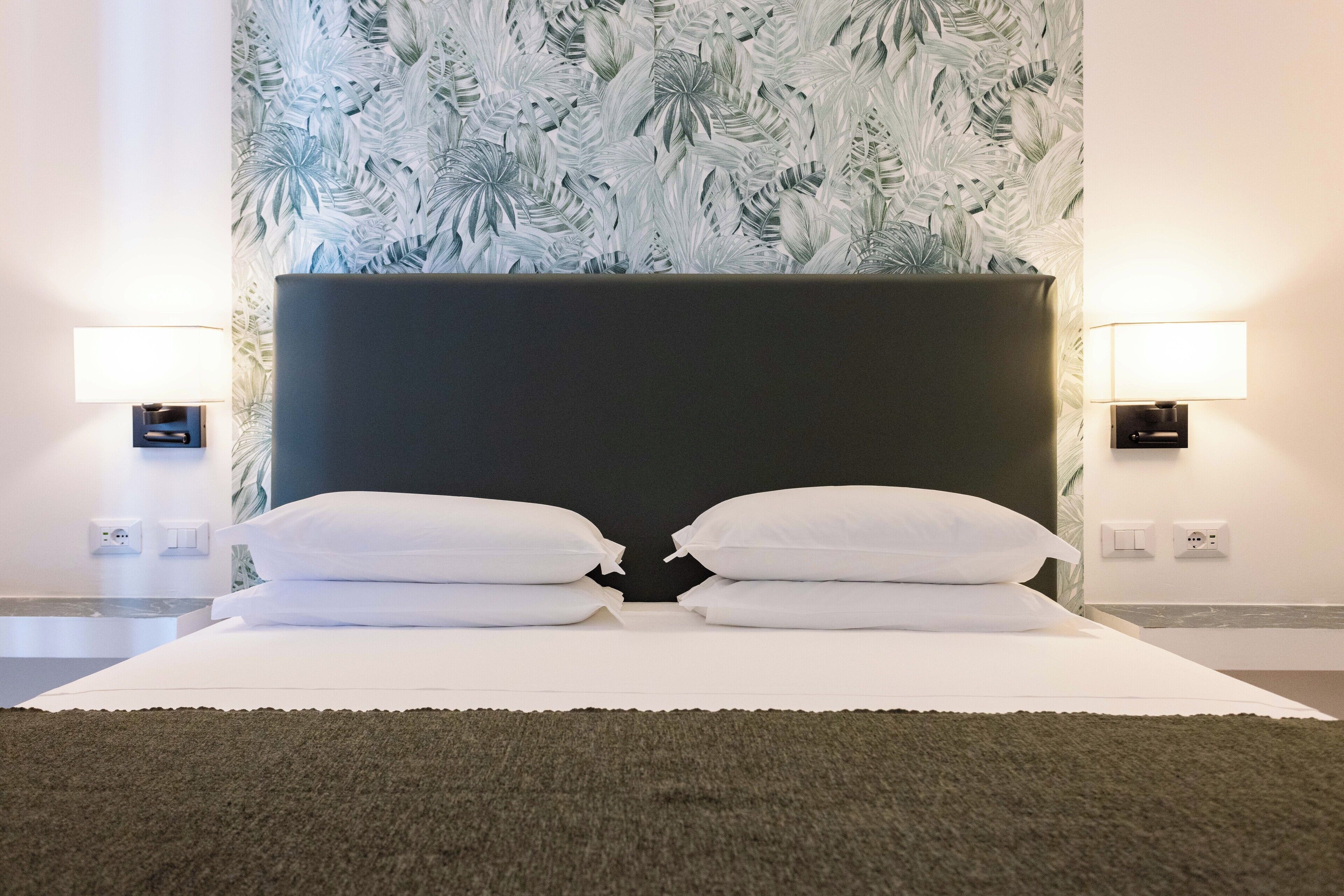 A hotel room with a large bed, two bedside lamps, and a tropical leaf-patterned wallpaper.