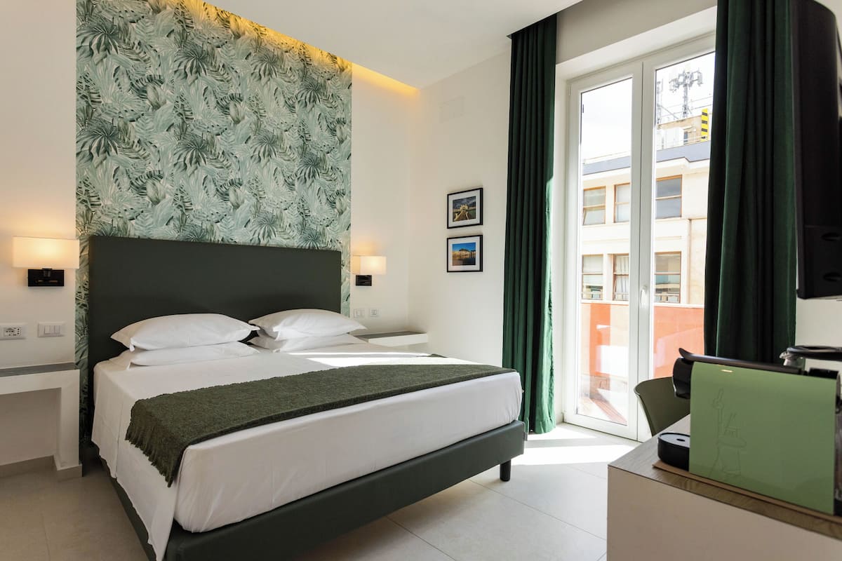 Ett hotellrum med en säng, ett skrivbord, en stol, ett fönster med gardiner och en vägg med ett lövmönster.