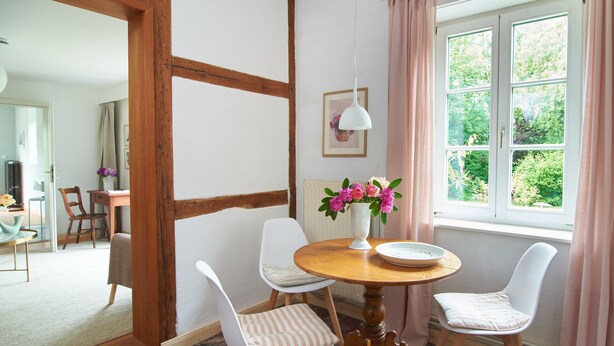 Un comedor con una mesa redonda de madera, cuatro sillas, un jarrón con flores y una ventana con cortinas.