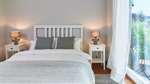 Un dormitorio con una cama, dos mesitas de noche con lámparas, un ventanal con cortinas y vista a la vegetación.