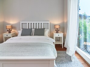 Ein Schlafzimmer mit einem Bett, zwei Nachttischen mit Lampen, einem Fenster mit Vorhängen und Blick ins Grüne.