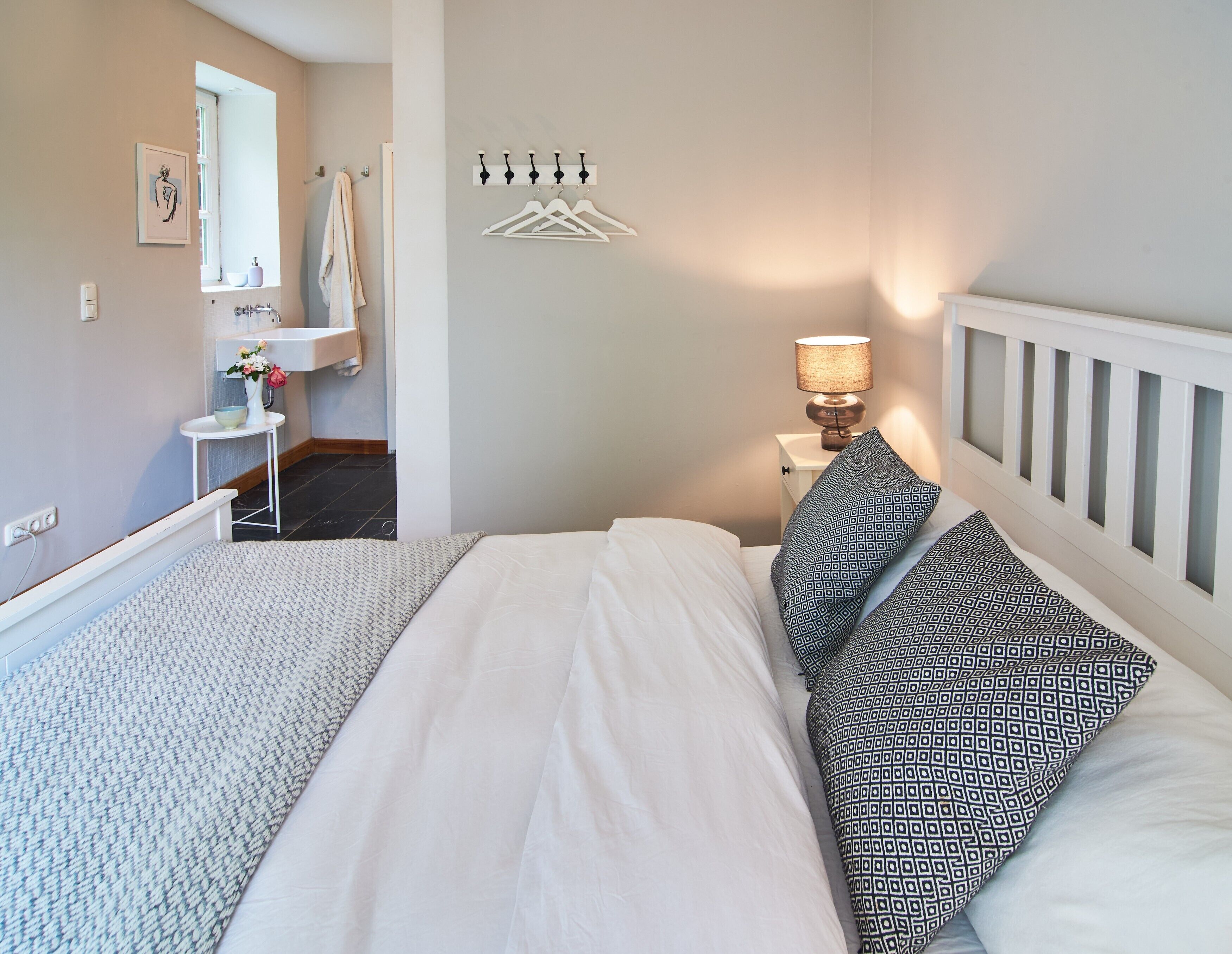 Ein Schlafzimmer mit Bett, Nachttisch mit Lampe, Kleiderhaken und einem Badezimmer mit Waschbecken und Handtuchhalter. Ein Fenster bietet einen Ausblick.