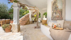A stone-floored patio with a wooden staircase, a seating area with cushions and a woven basket, and a wall-mounted painting. - Villa ARNELLA by Abahana Villas (Teulada)