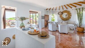 A spacious living area with a white sofa set, a coffee table, and a dining table with chairs. There are two windows providing natural light, and a large mirror hangs on the wall. - Villa ARNELLA by Abahana Villas (Teulada)
