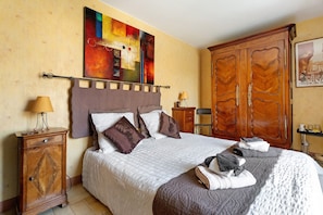 A hotel room with a large bed, a wooden wardrobe, a painting on the wall, and a small table with a lamp.