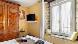Un dormitori amb un llit, un armari de fusta, una televisió sobre una estant, una finestra amb cortines i una paret de pedra.