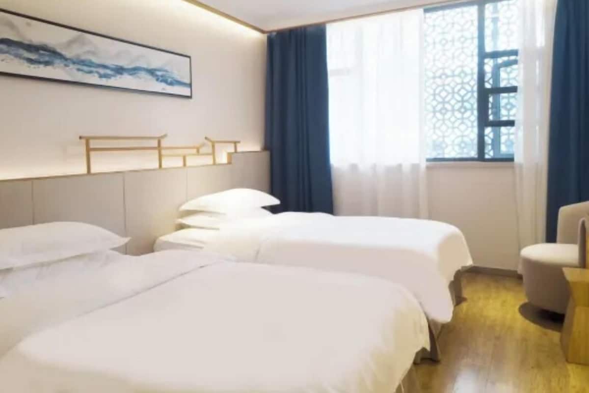 Quarto de hotel com duas camas, cabeceira de madeira, uma cadeira e uma janela com cortinas azuis.