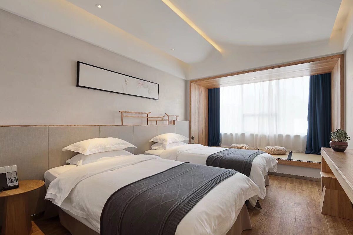 Quarto de hotel com duas camas, uma escrivaninha, janela com cortinas e um quadro na parede.