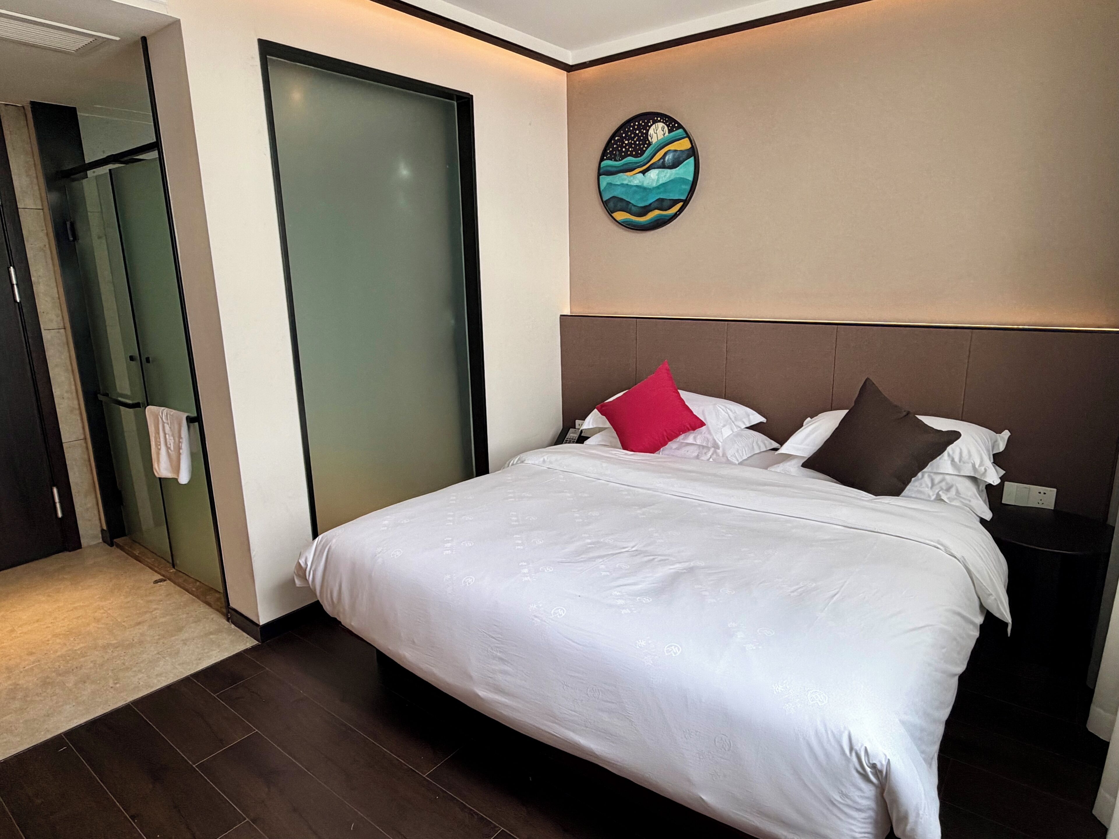 A hotel room with a bed, a nightstand, a painting, a door, and a bathroom.