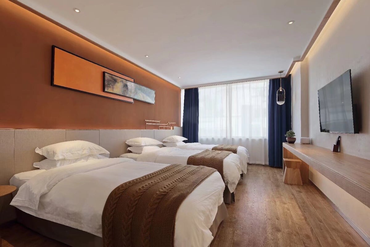 Quarto de hotel com três camas, uma televisão grande e uma escrivaninha de madeira.