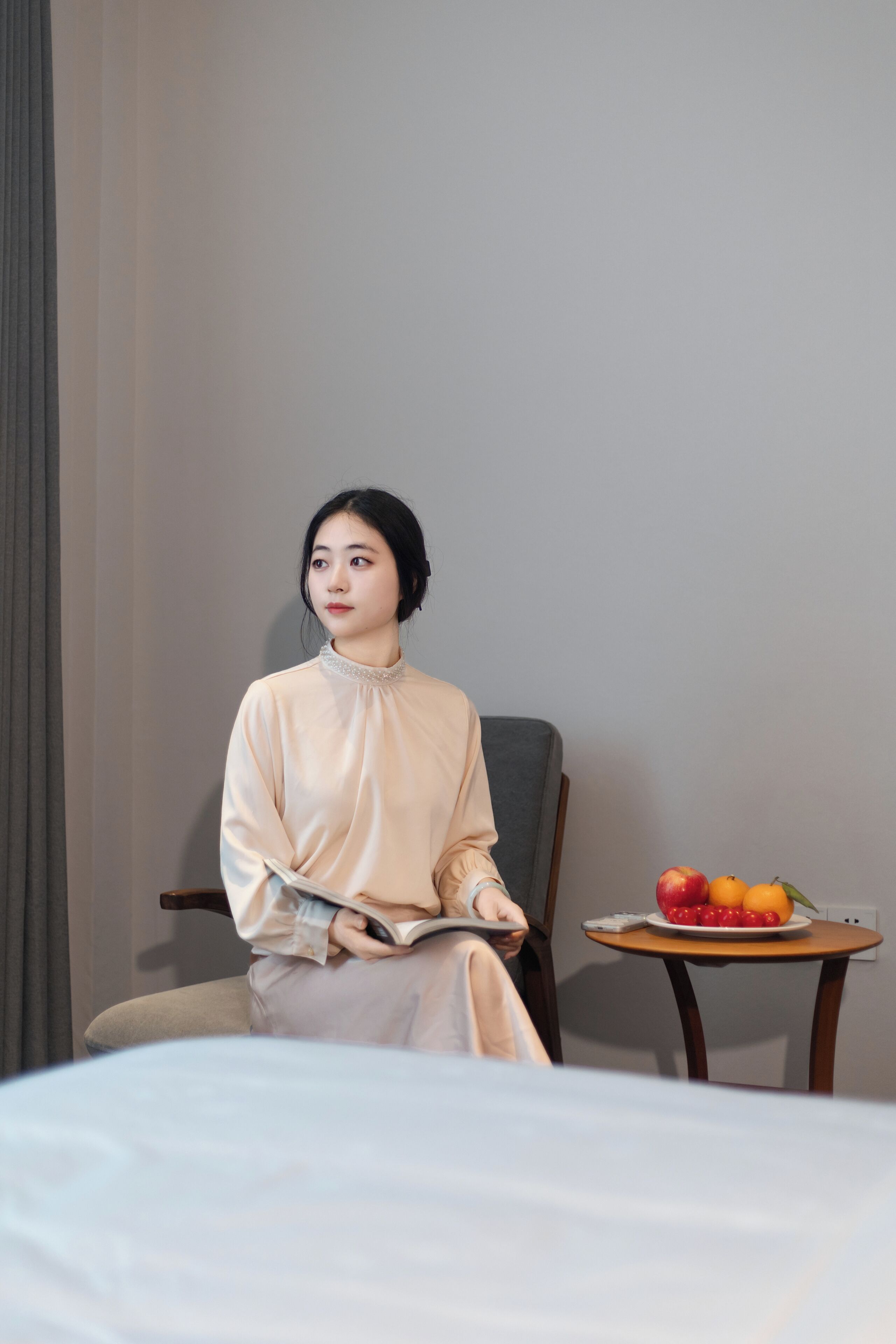 Una persona sentada en una habitación con una cama, una silla y una mesita con fruta y un libro.