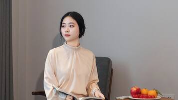 Una persona sentada en una habitación con una cama, una silla y una mesita con fruta y un libro.
