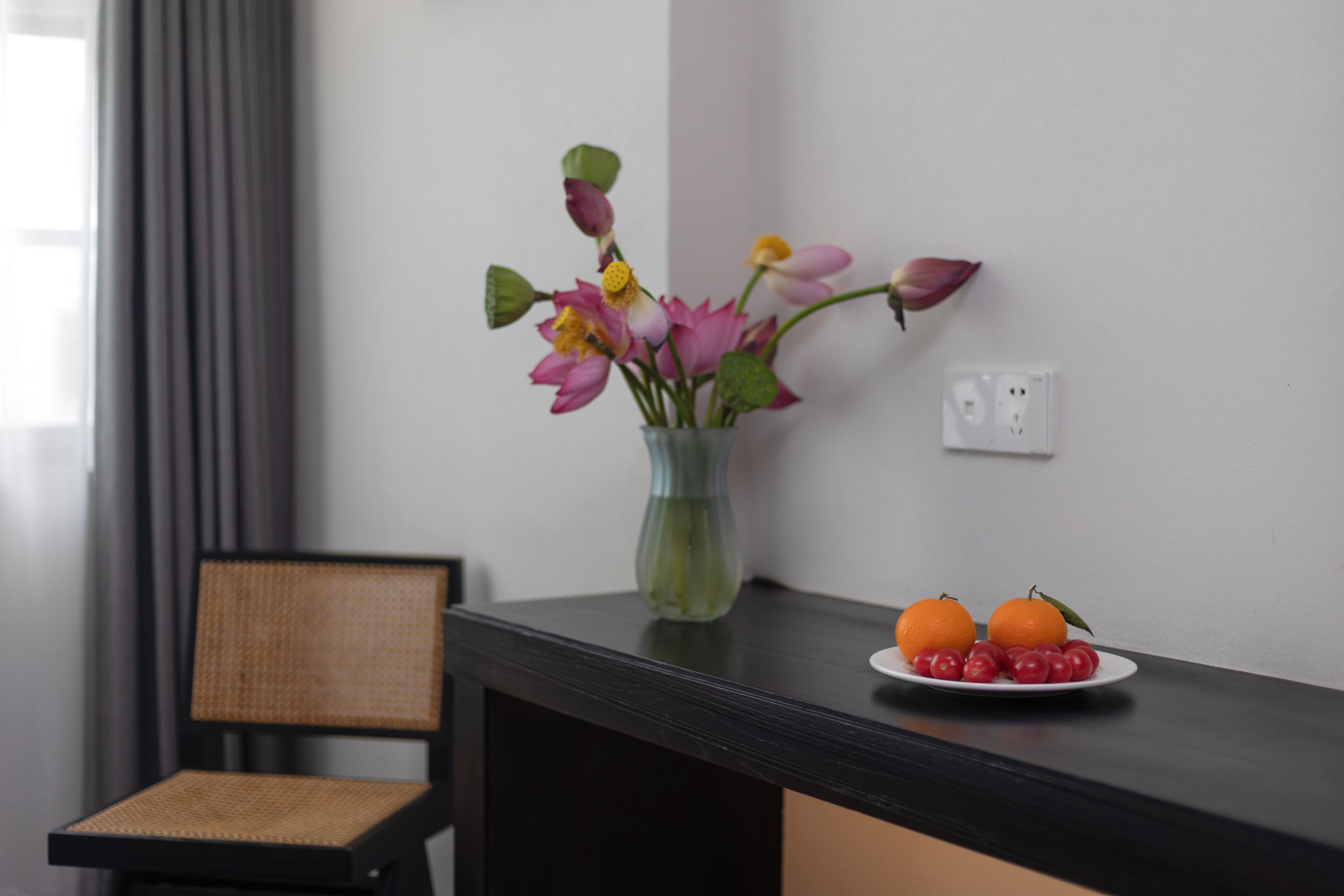 A room with a dark wooden table, a vase with flowers, a chair, and a plate with oranges and cherries.