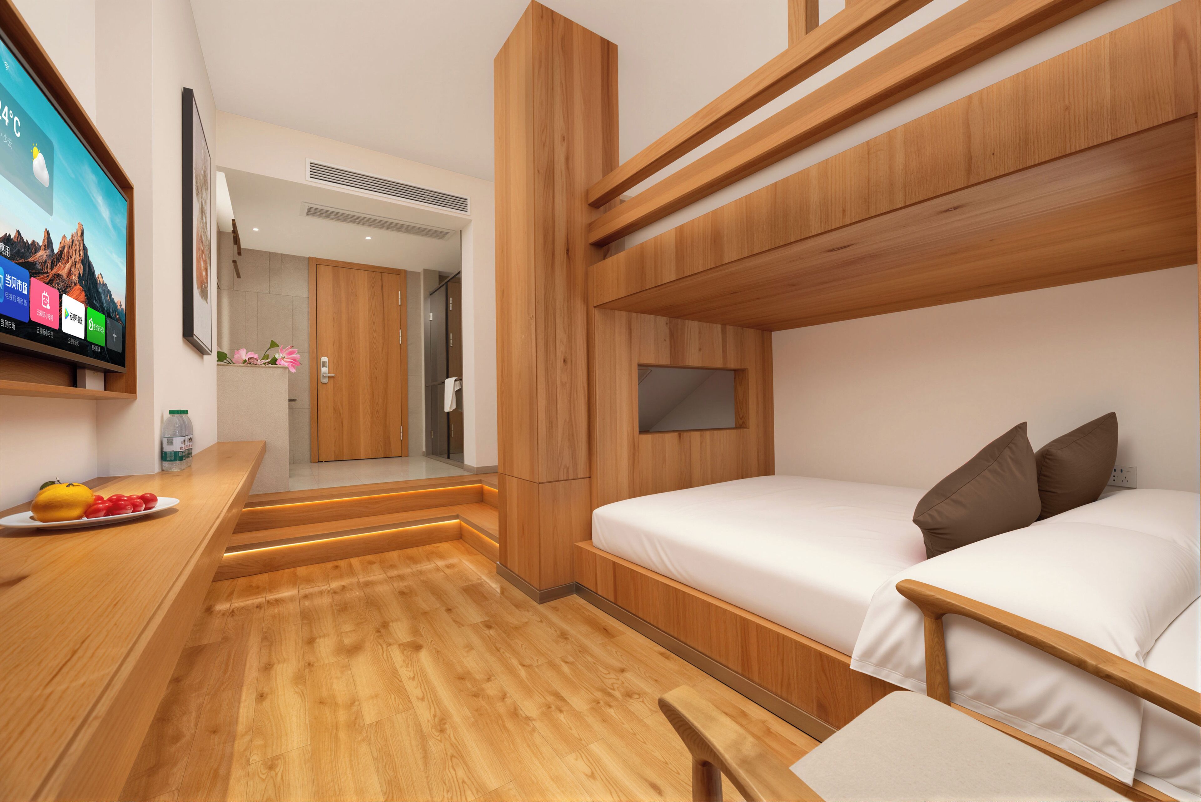 A modern hotel room with a wooden bunk bed, a flat-screen TV mounted on the wall, a small desk with a plate of fruit, and a bathroom visible through an open door.