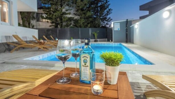 Escena a la piscina amb una taula de fusta, dues copes de vi, una ampolla de licor i una petita planta en test.