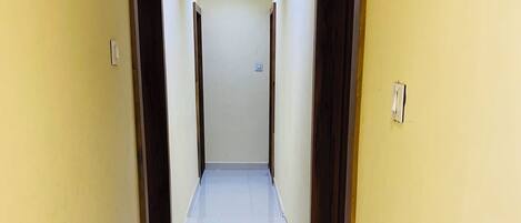 A narrow, well-lit hallway with yellow walls and a tiled floor, leading to a door at the end.