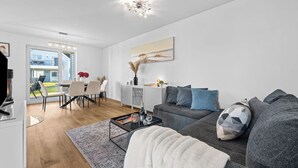 A modern living room with a grey sofa, a coffee table, and a dining area with a white table and chairs. - Ella - Terrace, 2 parking spaces, washing machine, fireplace, ground floor (Olpenitz)