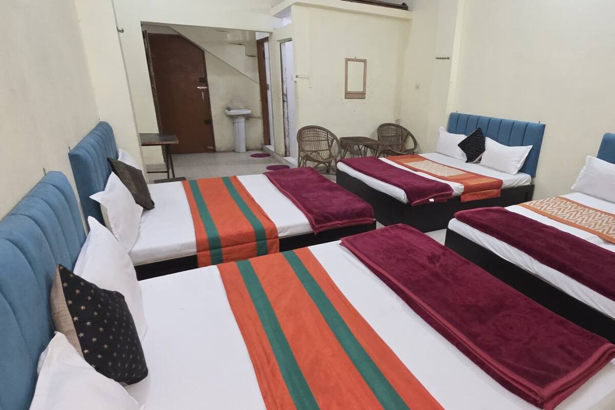 Une chambre d’hôtel avec trois lits, chacun doté d’une tête de lit de couleur différente et de plusieurs oreillers. On aperçoit une petite table et une chaise dans un coin, ainsi qu’un lavabo au fond de la pièce.