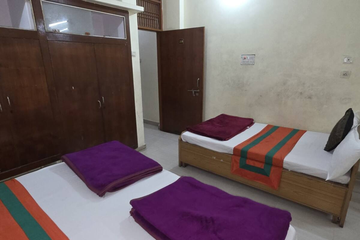 Une chambre avec une armoire en bois, un lit simple recouvert d’une couverture violette et orange, et une chaise en osier.